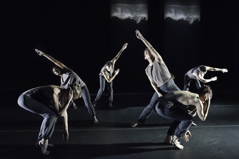 Six dancers arranged in a symmetrical configuration. Two dancers bend their torsos to rest on their thighs. Two dancers reach their arm that is center overhead on a diagonal. Two additional dancers upstage swing their arms to their sides. 